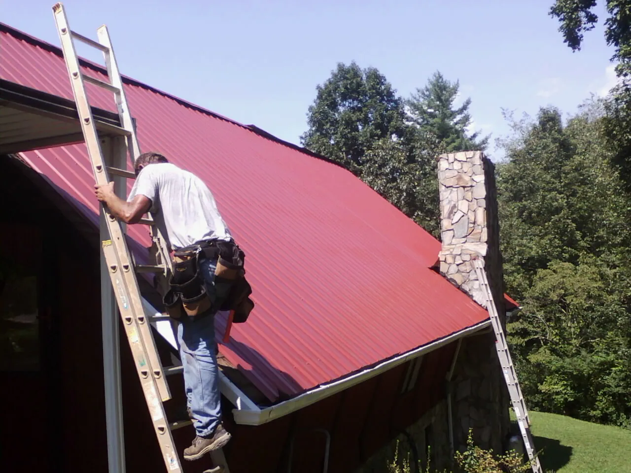 Roofer on a ladder beside a red metal roof and stone chimney — Soffit & Fascia Repair by Roofing Brothers in Beallsville