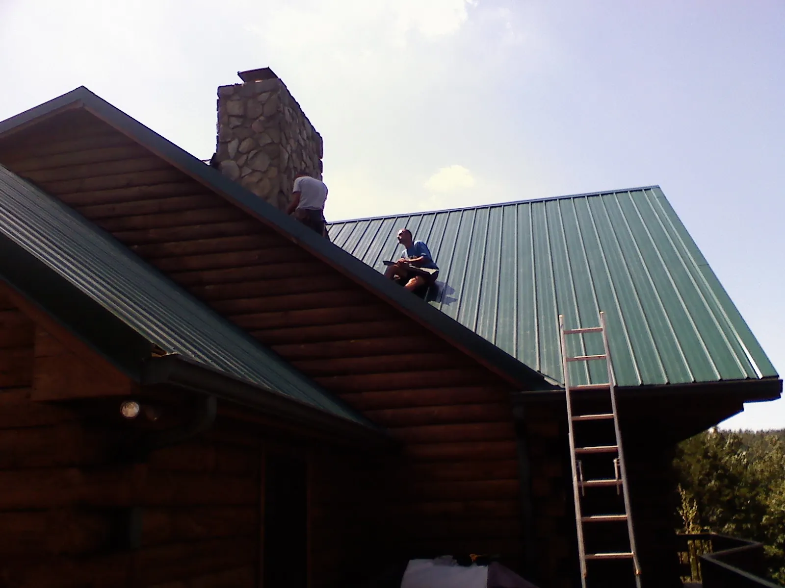 Roofers installing green metal panels beside a stone chimney on a log home in Beallsville